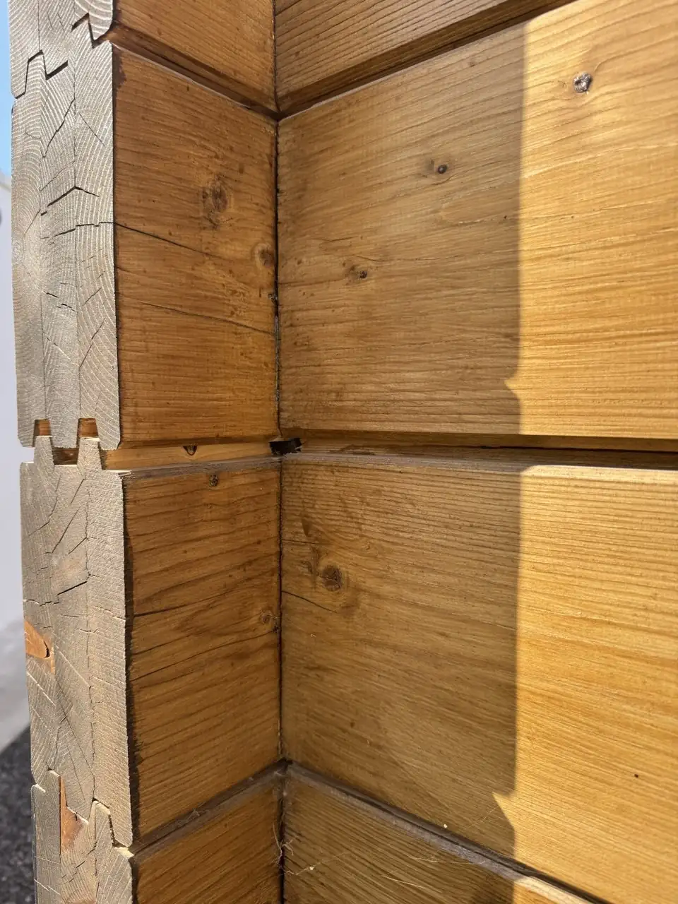 Esquina de una pared de madera con juntas horizontales visibles y grietas en los extremos de las piezas